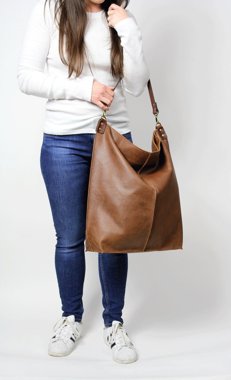 Puede incluir: Un bolso tote de cuero marr&oacute;n con una correa larga. El bolso es grande y espacioso, y lo lleva una persona que lleva jeans y una camisa blanca.