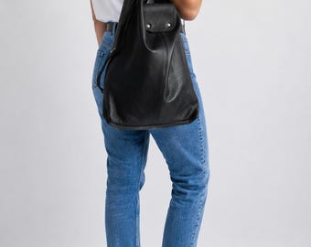May include: A black leather shoulder bag with a slouchy silhouette, featuring a fold-over flap with silver-toned hardware, a wide adjustable shoulder strap, and a spacious main compartment, worn over the shoulder against a white t-shirt and blue jeans.