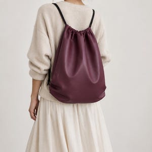 May include: A deep burgundy drawstring backpack made of a smooth, matte material. The bag features a gathered top closure with thin black cord straps that extend from the top corners to the base, providing a simple and functional design.