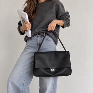 Può includere: Una borsa a spalla in pelle nera con silhouette rettangolare, patta ripiegabile e chiusura a girello color argento. La borsa presenta un unico cinturino sottile e una superficie testurizzata, offrendo un aspetto strutturato e definito.