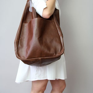 May include: A large brown leather tote bag with a single pocket on the front. The bag is worn over the shoulder of a person wearing a white shirt and skirt.