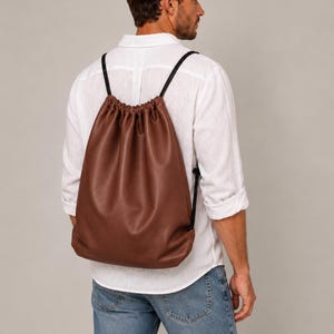 Peut inclure: Un sac à dos à cordon de couleur marron, fabriqué dans une matière lisse rappelant le cuir. Le sac présente une fermeture froncée sur le dessus avec de fines bretelles en cordon noir qui partent des côtés pour se rejoindre en haut.