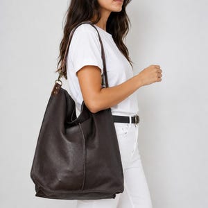 May include: A large, dark brown hobo-style shoulder bag crafted from textured leather. The bag features a slouchy, unstructured silhouette with a single wide strap attached by metal hardware, providing a spacious interior compartment.