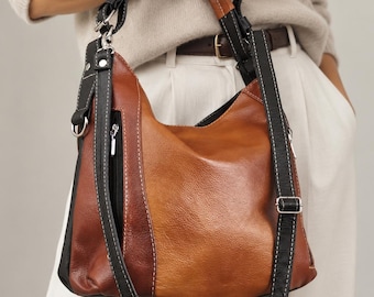 May include: A multi-toned brown leather hobo bag featuring a slouchy silhouette, a vertical zippered side pocket, and a long adjustable black leather shoulder strap with white contrast stitching, accented by silver-toned metal hardware and rings.