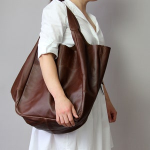 May include: A large brown leather tote bag with a slouchy design. The bag has a single strap and is being held by a person.