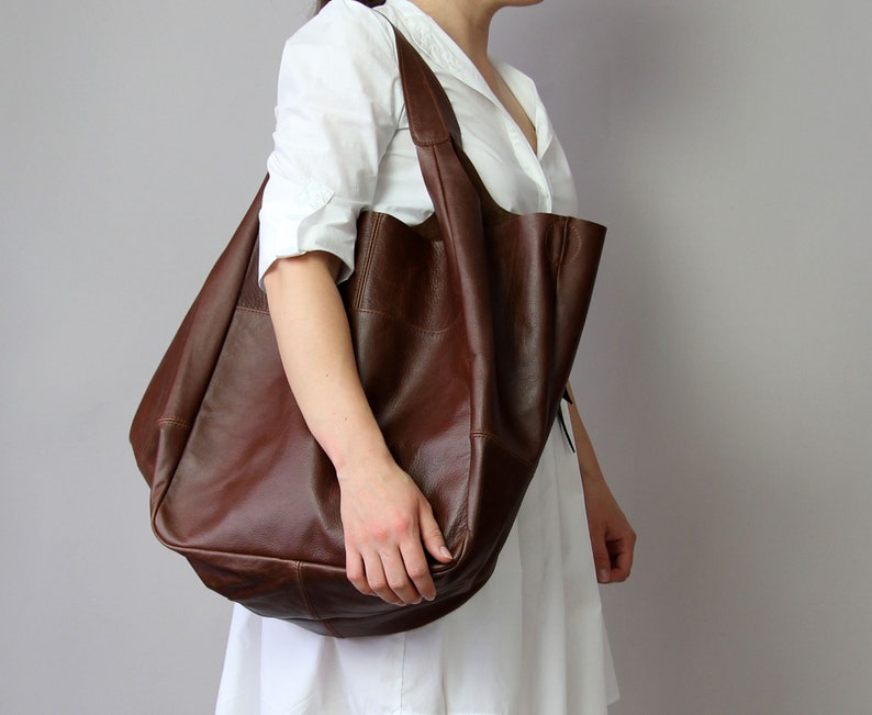 May include: A large brown leather tote bag with a slouchy design. The bag has a single strap and is being held by a person.