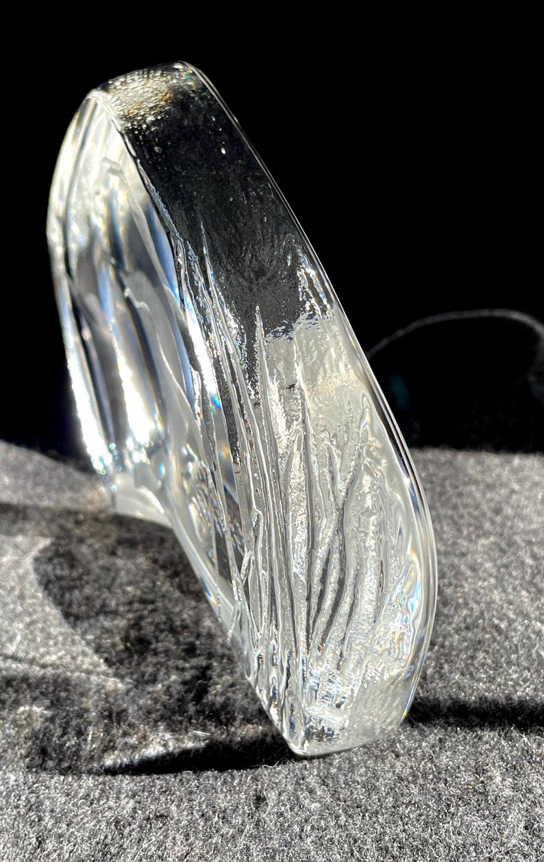 May include: A clear, sculpted glass object with a curved, organic shape. The glass has a textured surface with vertical lines and ridges, catching the light and creating a play of reflections. The object sits on a gray surface.