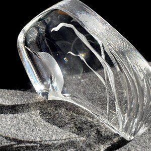 May include: A clear glass sculpture with a bird and plant design. The bird is etched into the glass, and the plants have a flowing, linear design. The sculpture is rectangular with rounded edges and sits on a gray surface.