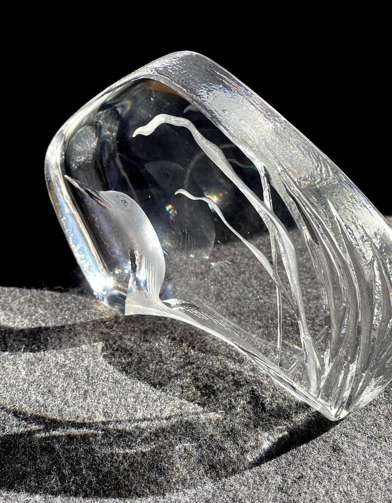 May include: A clear glass sculpture with a bird and plant design. The bird is etched into the glass, and the plants have a flowing, linear design. The sculpture is rectangular with rounded edges and sits on a gray surface.