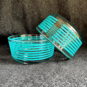 May include: Two clear glass bowls with turquoise horizontal stripes. One bowl is upright, and the other is tilted, resting against the first. The bowls have a retro, mid-century modern aesthetic.