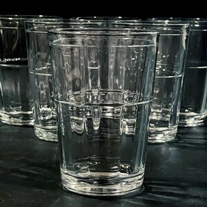 May include: A set of six clear, textured drinking glasses. The glasses are cylindrical with a grid-like pattern around the middle and a thick base. They are arranged in a semi-circle, with a dark background.