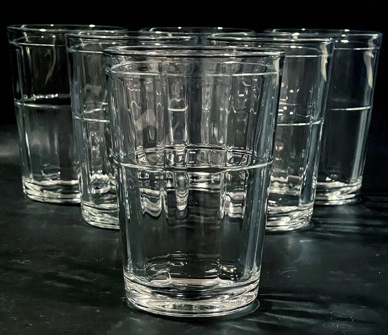 May include: A set of six clear, textured drinking glasses. The glasses are cylindrical with a grid-like pattern around the middle and a thick base. They are arranged in a semi-circle, with a dark background.