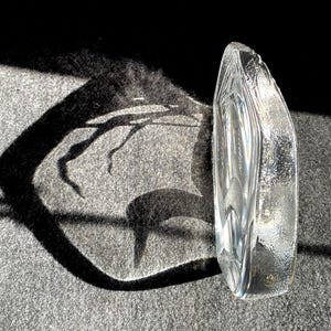 May include: A clear, textured glass sculpture with an organic, curved shape. The sculpture is positioned upright, catching the sunlight and casting a dark shadow on a gray surface. The glass has a textured surface and a smooth, polished interior.
