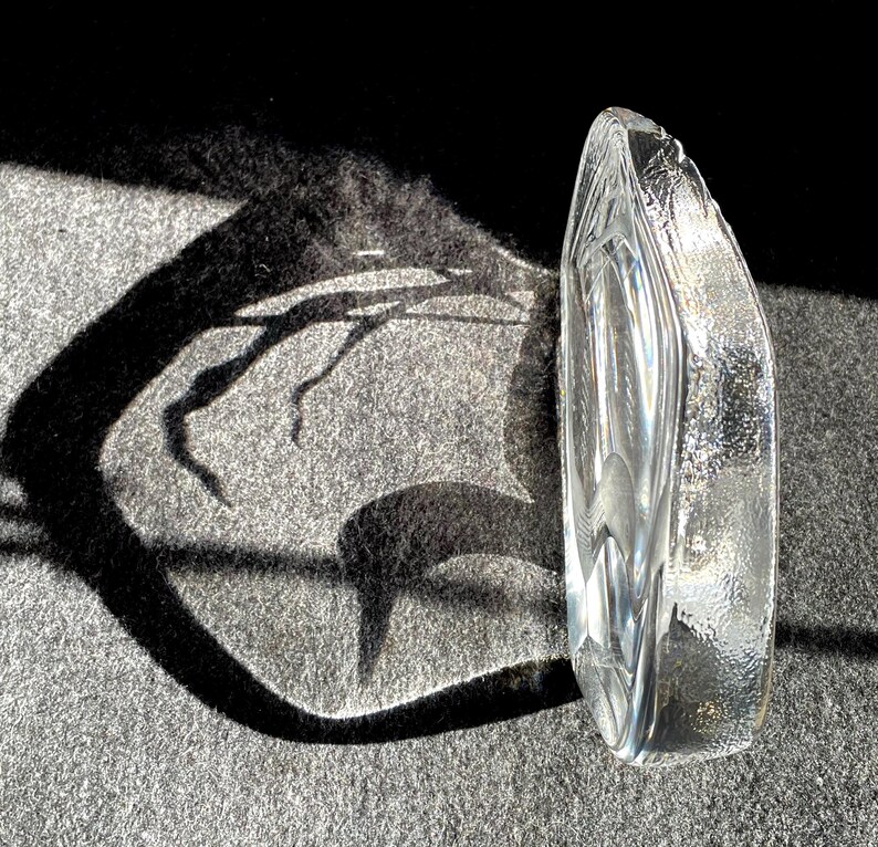 May include: A clear, textured glass sculpture with an organic, curved shape. The sculpture is positioned upright, catching the sunlight and casting a dark shadow on a gray surface. The glass has a textured surface and a smooth, polished interior.