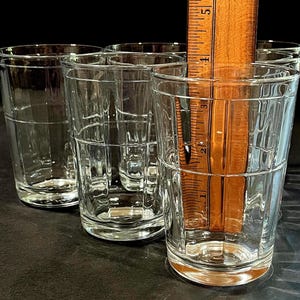 May include: A set of six clear, vintage-style drinking glasses. Each glass has a rectangular grid pattern and is approximately 4 inches tall. A wooden ruler is placed inside one glass for measurement. The glasses are on a dark surface.