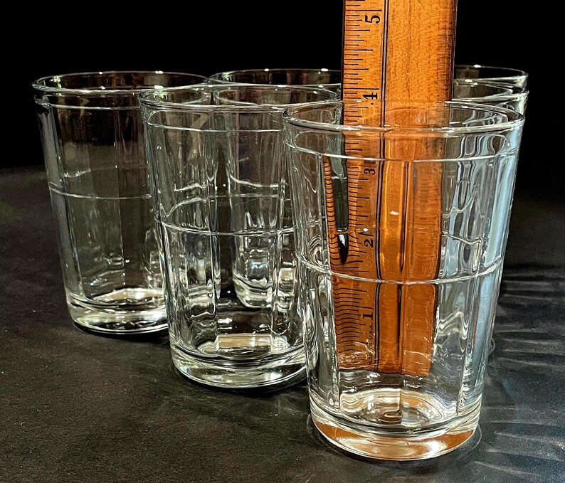 May include: A set of six clear, vintage-style drinking glasses. Each glass has a rectangular grid pattern and is approximately 4 inches tall. A wooden ruler is placed inside one glass for measurement. The glasses are on a dark surface.