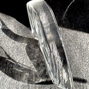 May include: A clear, curved glass object with a textured surface. The object is positioned at an angle, reflecting light and creating shadows. The overall shape is smooth and rounded, with a slightly rough texture.