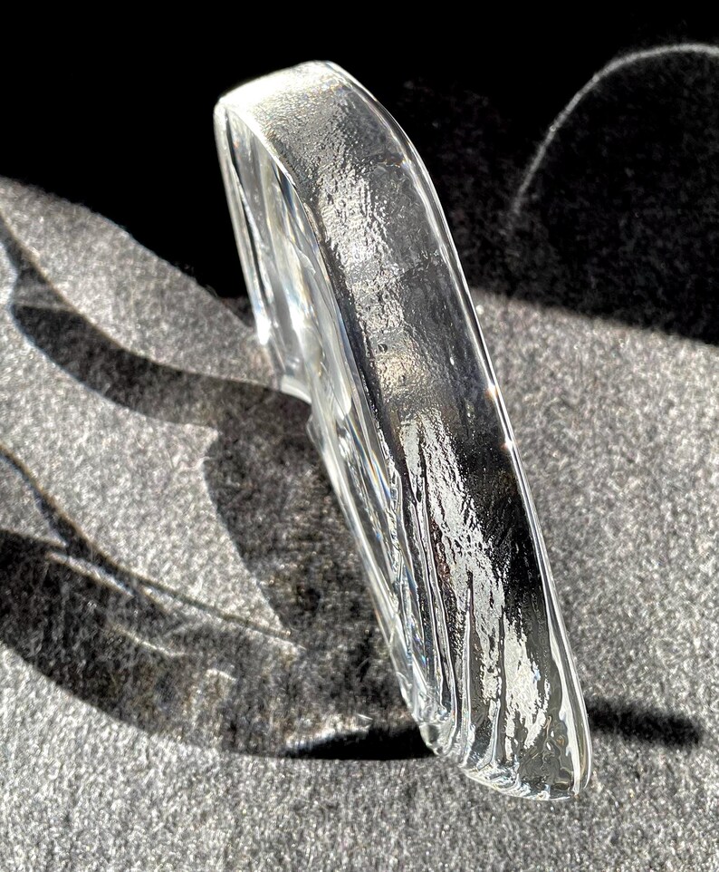 May include: A clear, curved glass object with a textured surface. The object is positioned at an angle, reflecting light and creating shadows. The overall shape is smooth and rounded, with a slightly rough texture.