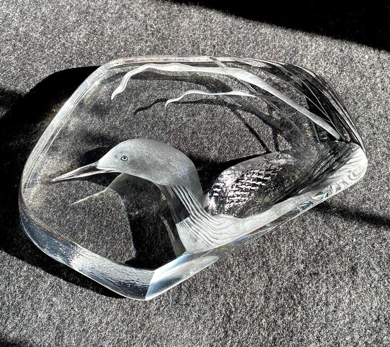May include: A clear, glass sculpture of a bird, possibly a loon, with etched details. The bird is within a clear, rectangular frame with rounded edges. The bird's head and body are detailed with etched lines and textures.
