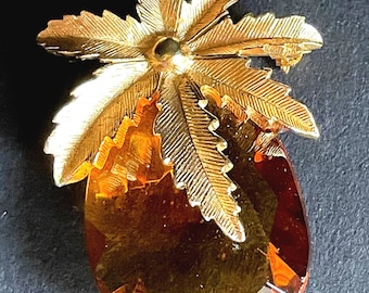 Op de afbeelding: Een broche van goudkleurig metaal met een gestileerde palmboom en een grote, amberkleurige glazen steen. De steen is gefacetteerd en heeft een licht ovale vorm.
