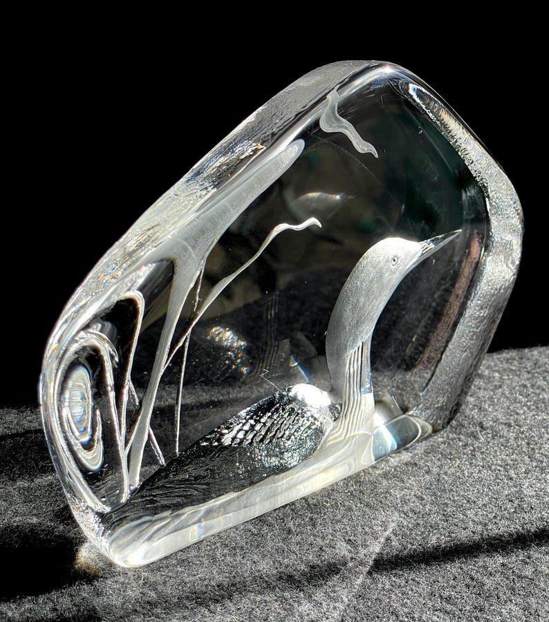 May include: A clear glass sculpture of a bird, possibly a loon, with etched details. The bird has a gray head and black and white body. The sculpture is rectangular with rounded edges and sits on a gray surface.