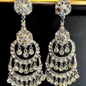 May include: A pair of silver dangle earrings with multiple tiers of clear crystal stones. The earrings have a circular design with a large crystal stone in the center.
