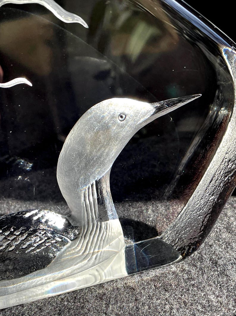 May include: A clear glass sculpture of a bird with a long, slender beak and detailed feather etching. The bird's head and neck are prominently displayed, with a textured surface and a curved shape. The sculpture is set against a dark background.