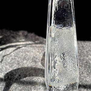 May include: A clear, rectangular glass vase with a textured surface. The vase is slightly tapered and stands upright, casting a shadow on a gray surface. The glass has a frosted appearance.