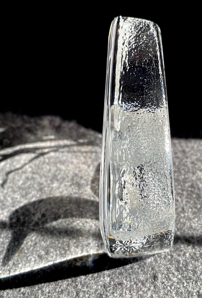 May include: A clear, rectangular glass vase with a textured surface. The vase is slightly tapered and stands upright, casting a shadow on a gray surface. The glass has a frosted appearance.