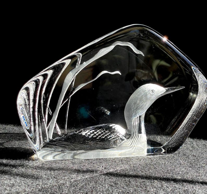 May include: A clear glass sculpture of a loon bird, with etched details and a stylized, abstract design. The bird is depicted with a long neck and beak, set within a clear, rectangular frame. The sculpture is on a gray surface.