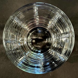 May include: A clear, textured glass with a circular design. The glass has a series of concentric rings and vertical lines, creating a grid-like pattern. The glass is empty and reflects the surrounding light, creating a play of light and shadow.