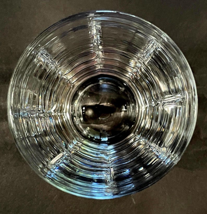 May include: A clear, textured glass with a circular design. The glass has a series of concentric rings and vertical lines, creating a grid-like pattern. The glass is empty and reflects the surrounding light, creating a play of light and shadow.