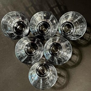 May include: Six clear, round drinking glasses arranged in a circular pattern. Each glass has a textured, ribbed design on the exterior. The glasses are empty, and the bottoms are visible, reflecting the dark surface below.