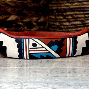Puede incluir: Un cuenco decorativo con forma de canoa pintado a mano. El cuenco es principalmente rojo con diseños geométricos blancos, negros, azules y marrones. El borde es rojo y el interior no está pintado. El cuenco está sobre una superficie clara.
