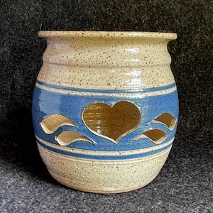 May include: A ceramic vessel with a speckled cream glaze and a wide blue horizontal band around the center. The blue section features a large heart-shaped cutout in the middle, flanked by two smaller leaf-shaped cutouts on each side of the heart.
