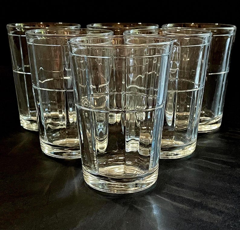 May include: Six clear, textured drinking glasses arranged in a group. The glasses have a rectangular grid pattern and a simple, elegant design. The glasses are empty and appear to be made of glass.