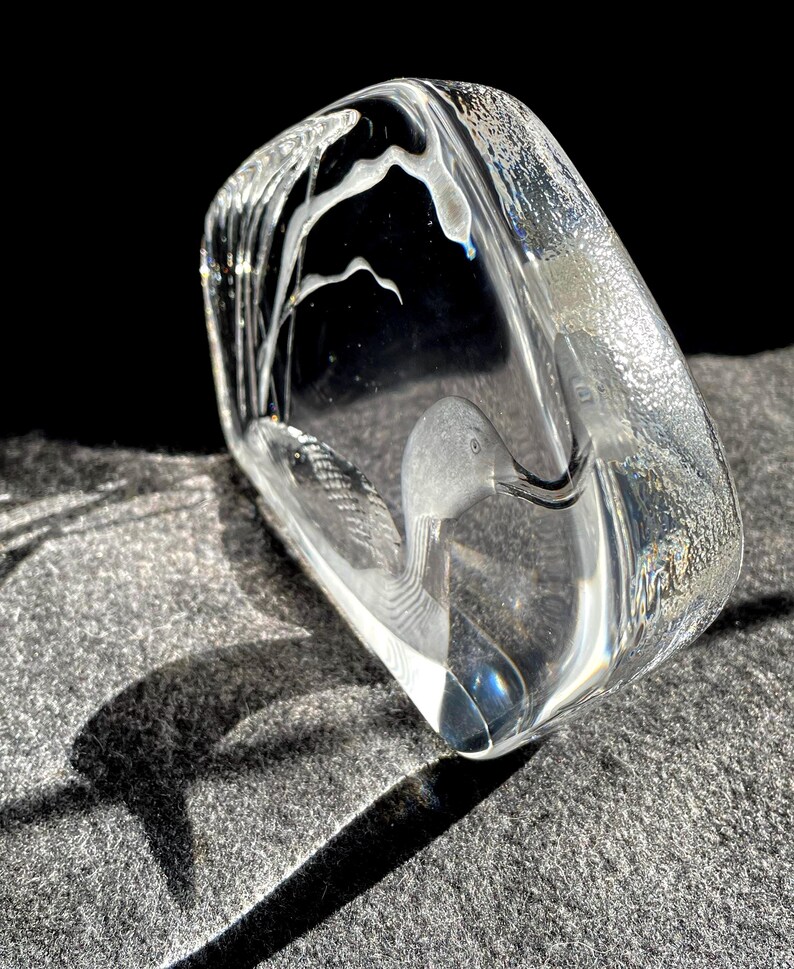 May include: A clear glass sculpture of a duck with etched details. The duck is inside a rectangular block of glass, with a textured surface and a dark background. The sculpture is illuminated, creating shadows.