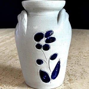 May include: A white ceramic vase with a textured surface and two handles. The vase features a dark blue floral design with a flower and leaves. The vase is a decorative item.
