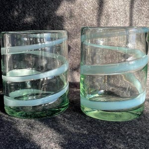 May include: Two clear, cylindrical drinking glasses with a green tint. Each glass features a light blue spiral design wrapping around the exterior. The glasses are made of glass and are sitting on a dark surface.