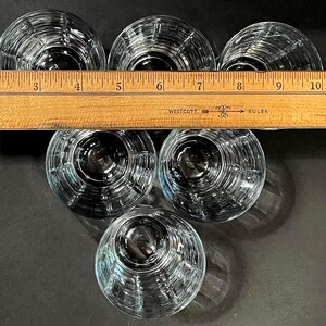 May include: Six clear, round drinking glasses with a ribbed design are arranged in a circular pattern. A wooden ruler with inch markings is placed across the top of the glasses. The glasses are on a dark surface.