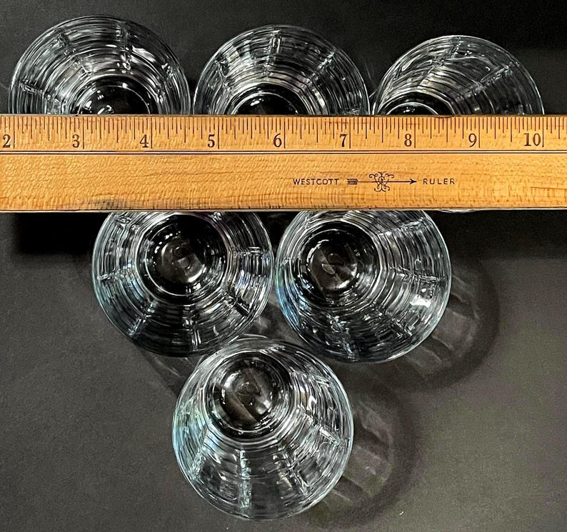 May include: Six clear, round drinking glasses with a ribbed design are arranged in a circular pattern. A wooden ruler with inch markings is placed across the top of the glasses. The glasses are on a dark surface.