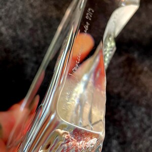 May include: A clear, rectangular glass object with a textured surface. The glass has a slight curve and appears to be hand-blown. The object has etched text on the side. The background is a dark, textured fabric.