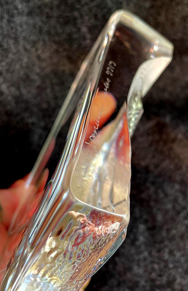 May include: A clear, rectangular glass object with a textured surface. The glass has a slight curve and appears to be hand-blown. The object has etched text on the side. The background is a dark, textured fabric.