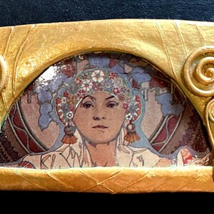 May include: A gold-coloured frame with a decorative scroll design surrounds a vintage image of a woman wearing a floral headdress. The woman's face is visible, and she is looking directly at the viewer.