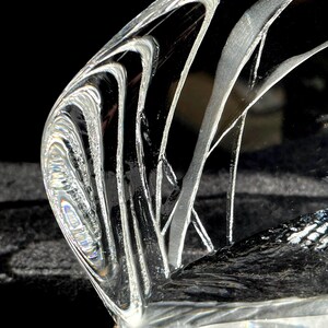 May include: A clear, curved glass object with etched, flowing white lines. The glass has a polished, reflective surface, and the design creates a sense of movement and depth. The object is set against a dark background.
