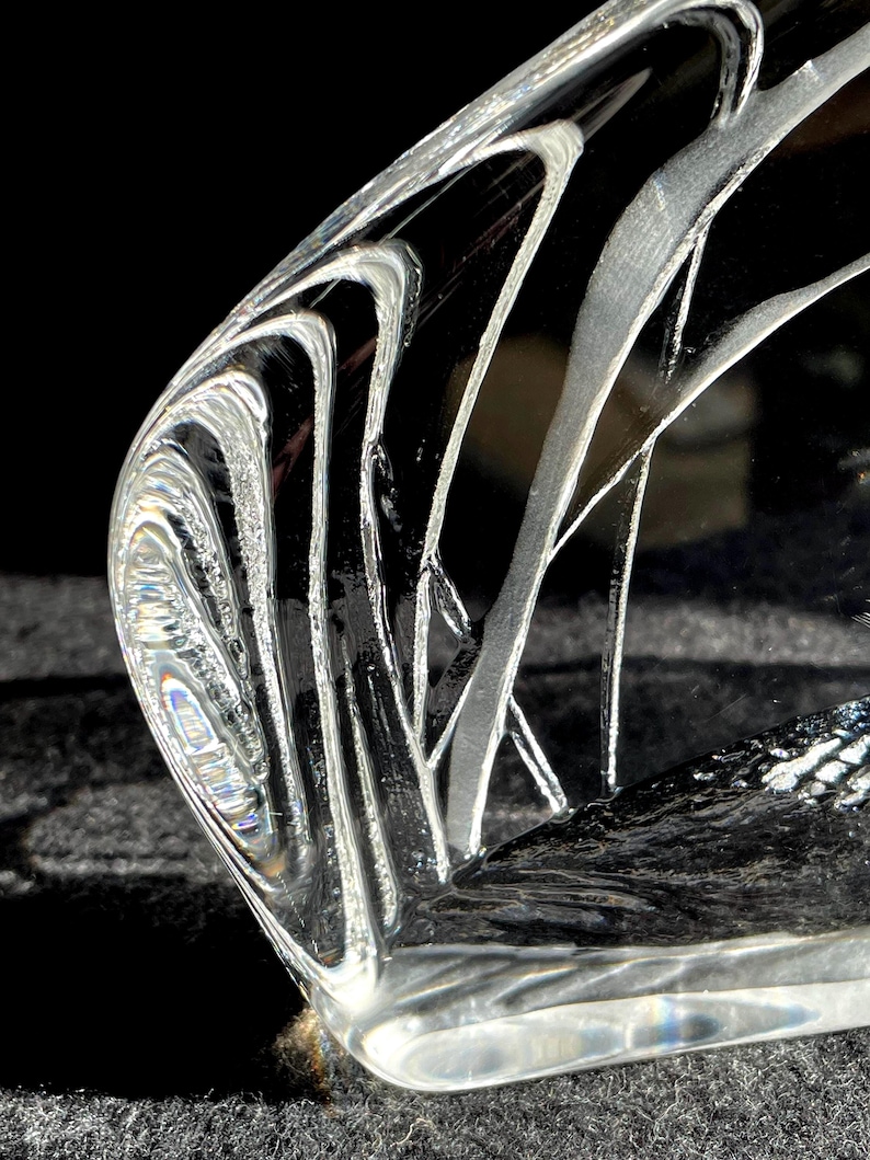 May include: A clear, curved glass object with etched, flowing white lines. The glass has a polished, reflective surface, and the design creates a sense of movement and depth. The object is set against a dark background.