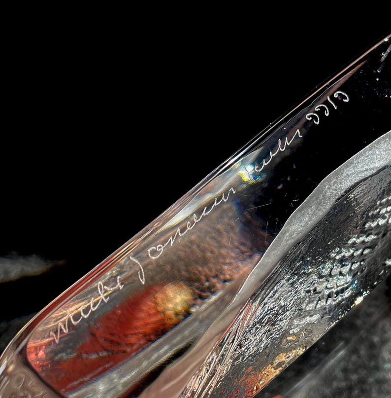 May include: Close-up of a clear, angled glass sculpture with etched white text. The sculpture has a textured surface and reflects light, with hints of red and yellow visible within. The text appears to be a signature and date.