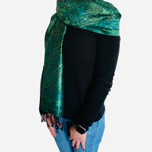 May include: A green paisley patterned scarf with a fringed edge. The scarf is draped around a person's neck and shoulders.