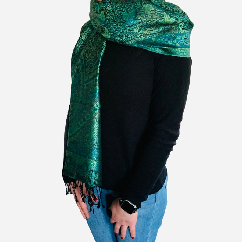 May include: A green paisley patterned scarf with a fringed edge. The scarf is draped around a person's neck and shoulders.