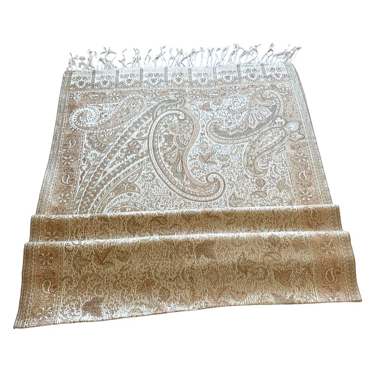 May include: A beige paisley patterned shawl with a fringed edge. The shawl is folded in half and the fringe is visible on the bottom edge.